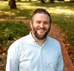 A man with a beard and a content smile is standing outdoors. He is wearing a light blue, long-sleeved button-up shirt and is posing for the camera with a relaxed, casual stance. The background is softly blurred, highlighting a park-like setting with greenery and trees that suggest it might be autumn, given the presence of fallen leaves on the ground. The lighting is warm, indicating the photo may have been taken on a sunny day. Photo credit: Kelly Darree @Kelly Darree Photography