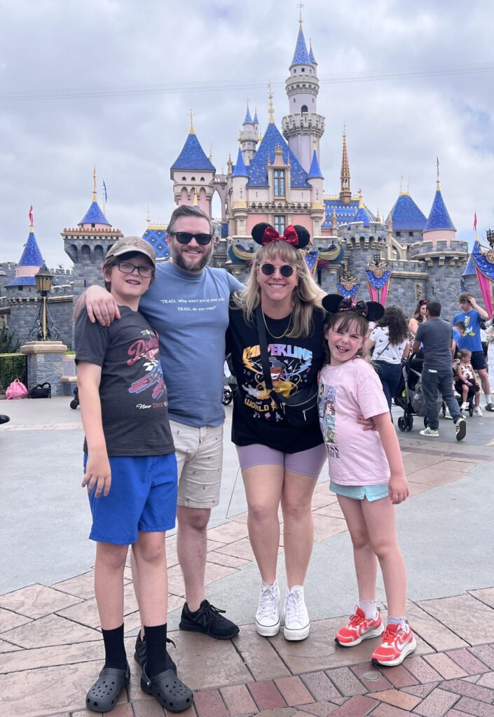Alexander Swan stands with his family in front of Sleeping Beauty Castle at Disneyland. Everyone is smiling and wearing Disney-themed attire, including Mickey ears on two family members, with the castle and visitors filling the background under a cloudy sky.